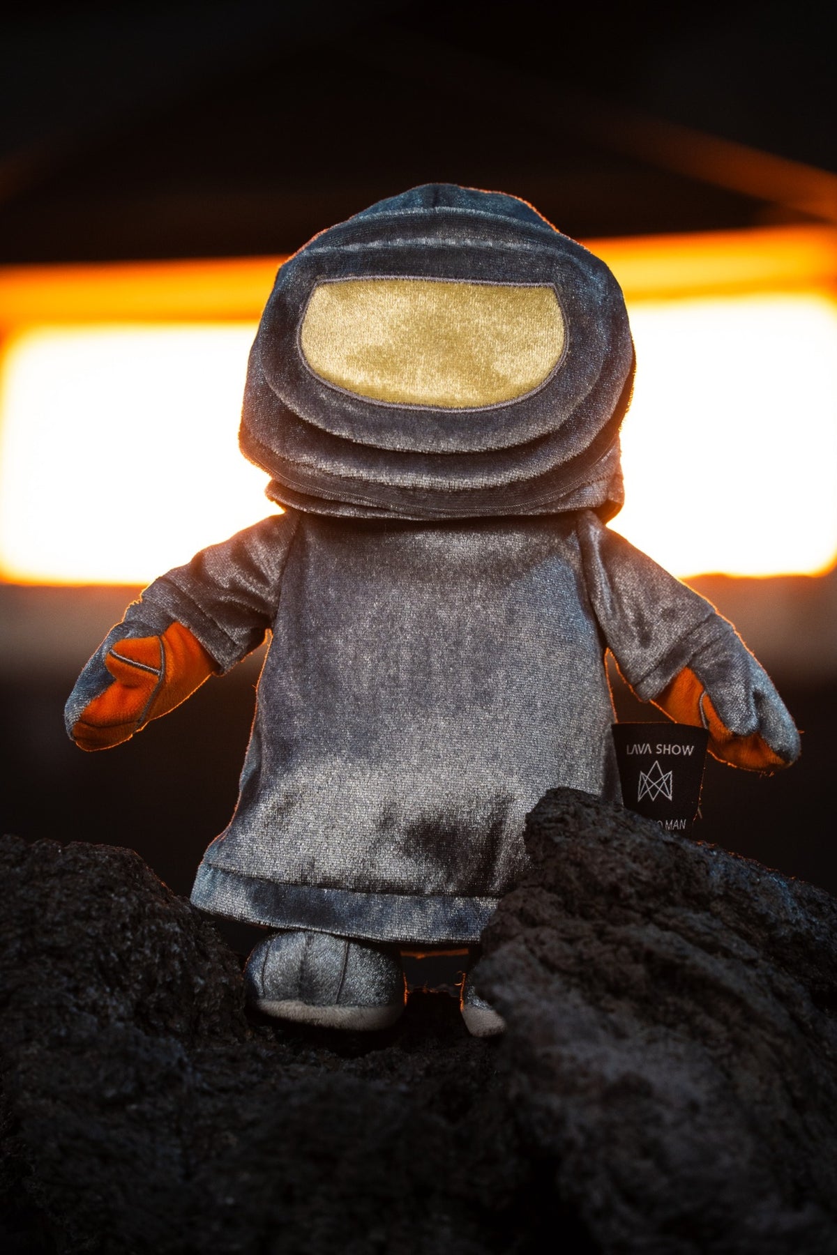 A plush Lava Man plushie standing on dark volcanic rock with glowing lava behind it. The toy is made of textured gray fabric with orange gloves and a gold visor.