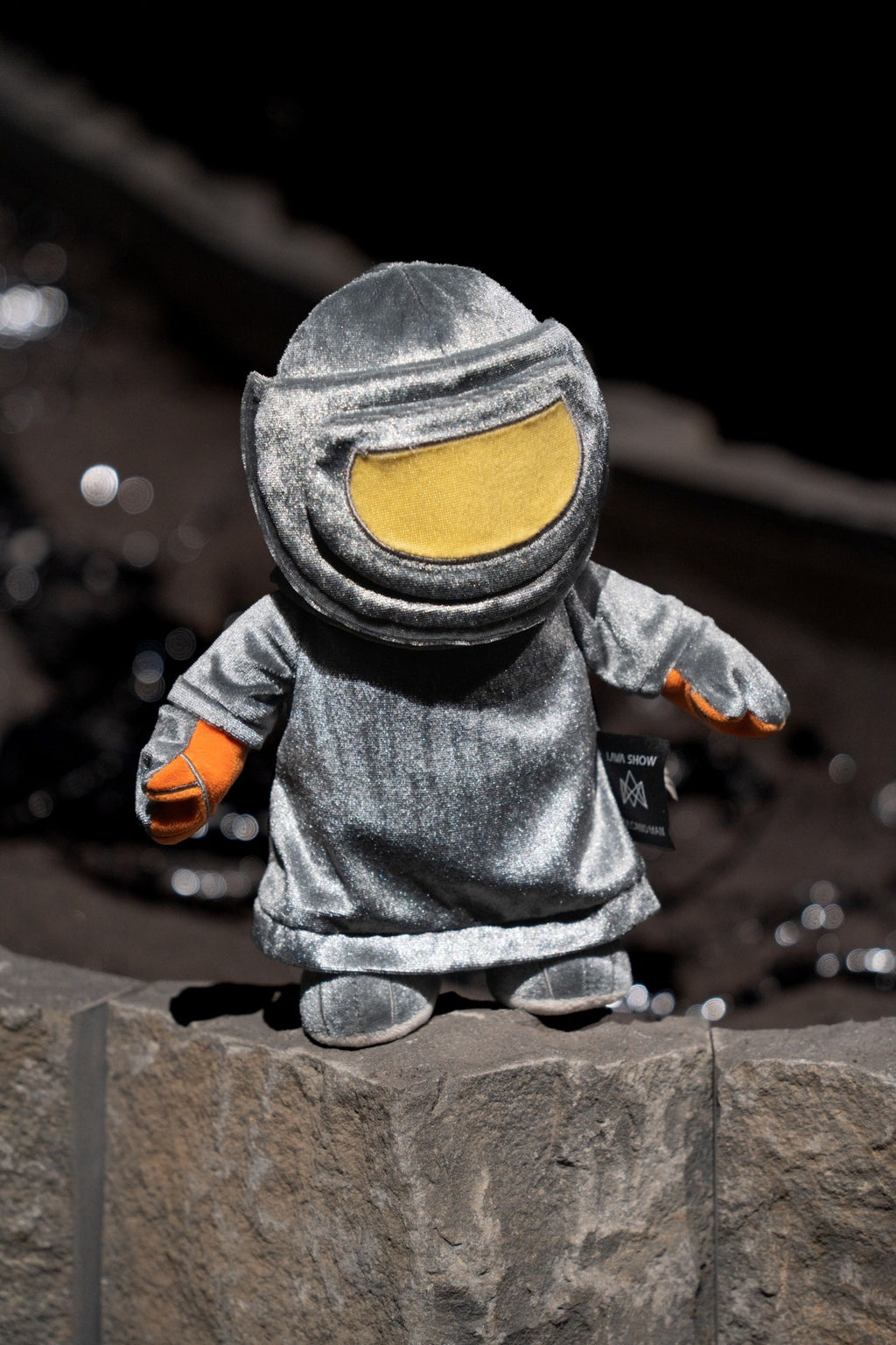 A small plush toy dressed in a metallic heat protective suit with a large yellow visor and orange gloves, standing on a stone surface against a dark blurred background.