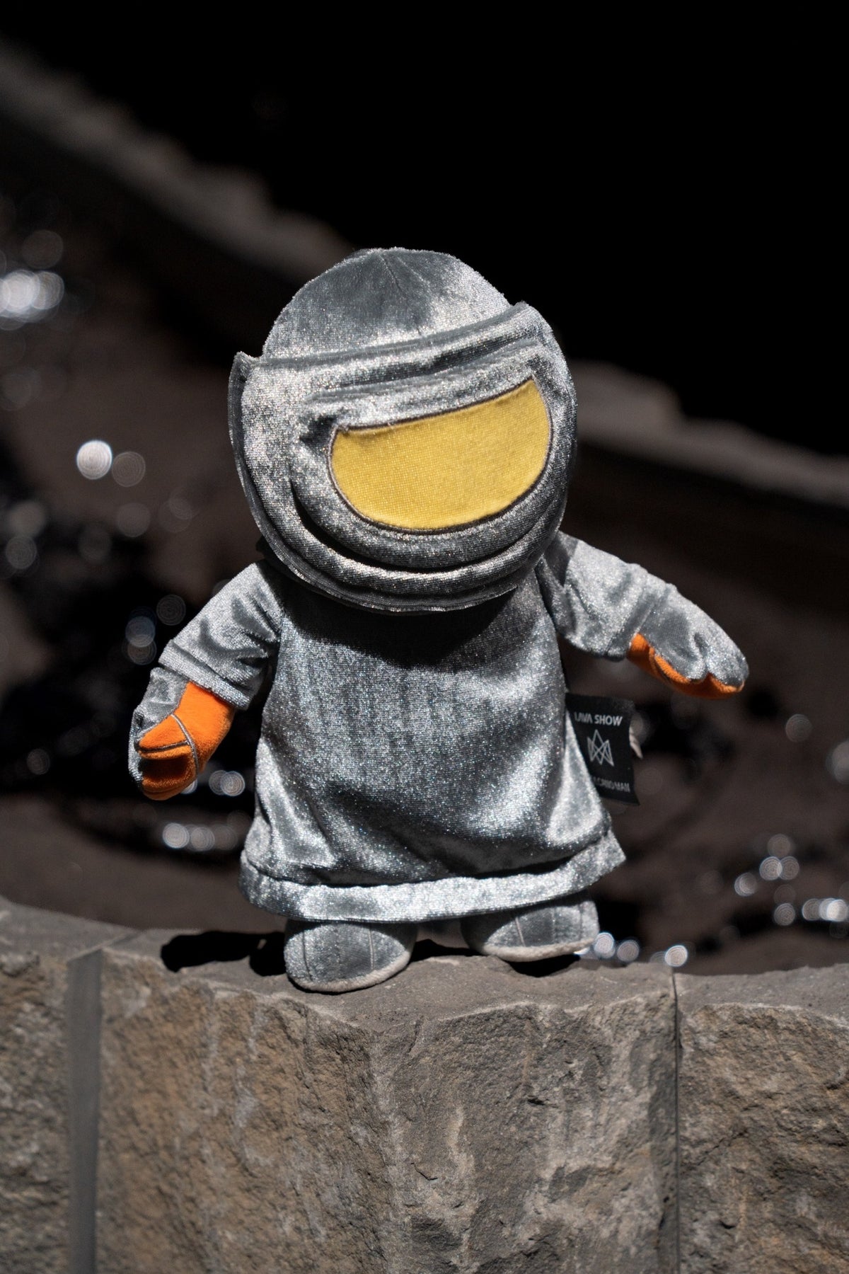 A small plush toy dressed in a metallic heat protective suit with a large yellow visor and orange gloves, standing on a stone surface against a dark blurred background.