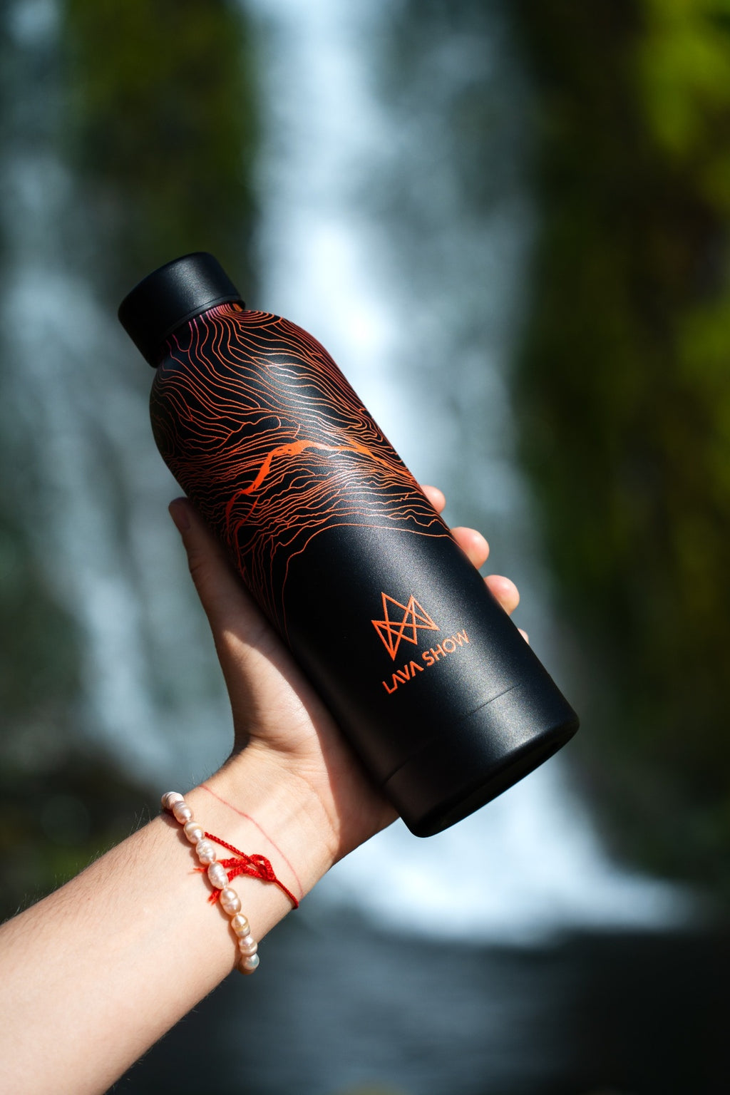 Black 500 ml Lava Show water bottle with orange flowing lava contour graphics and logo, held in one hand in front of a waterfall.