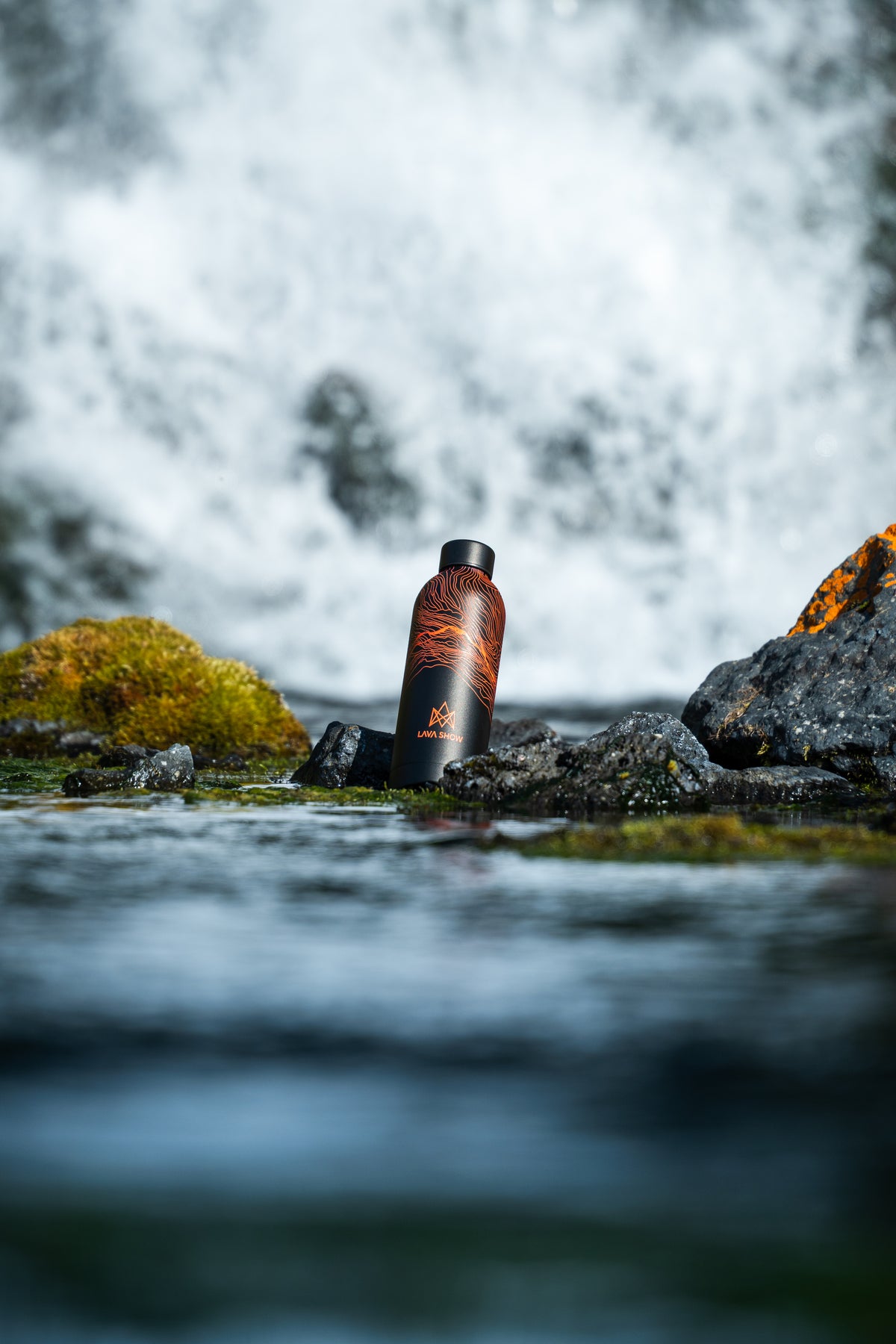 Black 500 ml Lava Show water bottle with orange lava contour graphics, standing among moss-covered rocks in front of a waterfall.